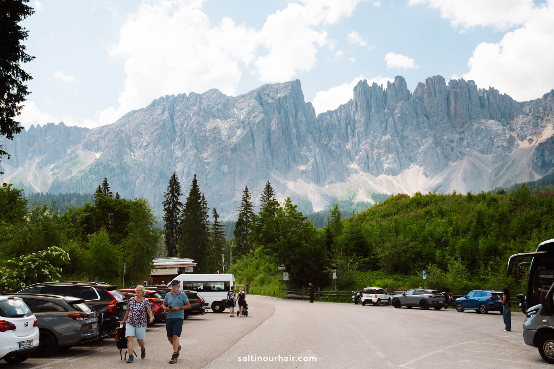 A parking lot with several cars and people, surrounded by green trees, with tall jagged mountains in the background under a partly cloudy sky—an ideal starting point for exploring things to do in the Dolomites.