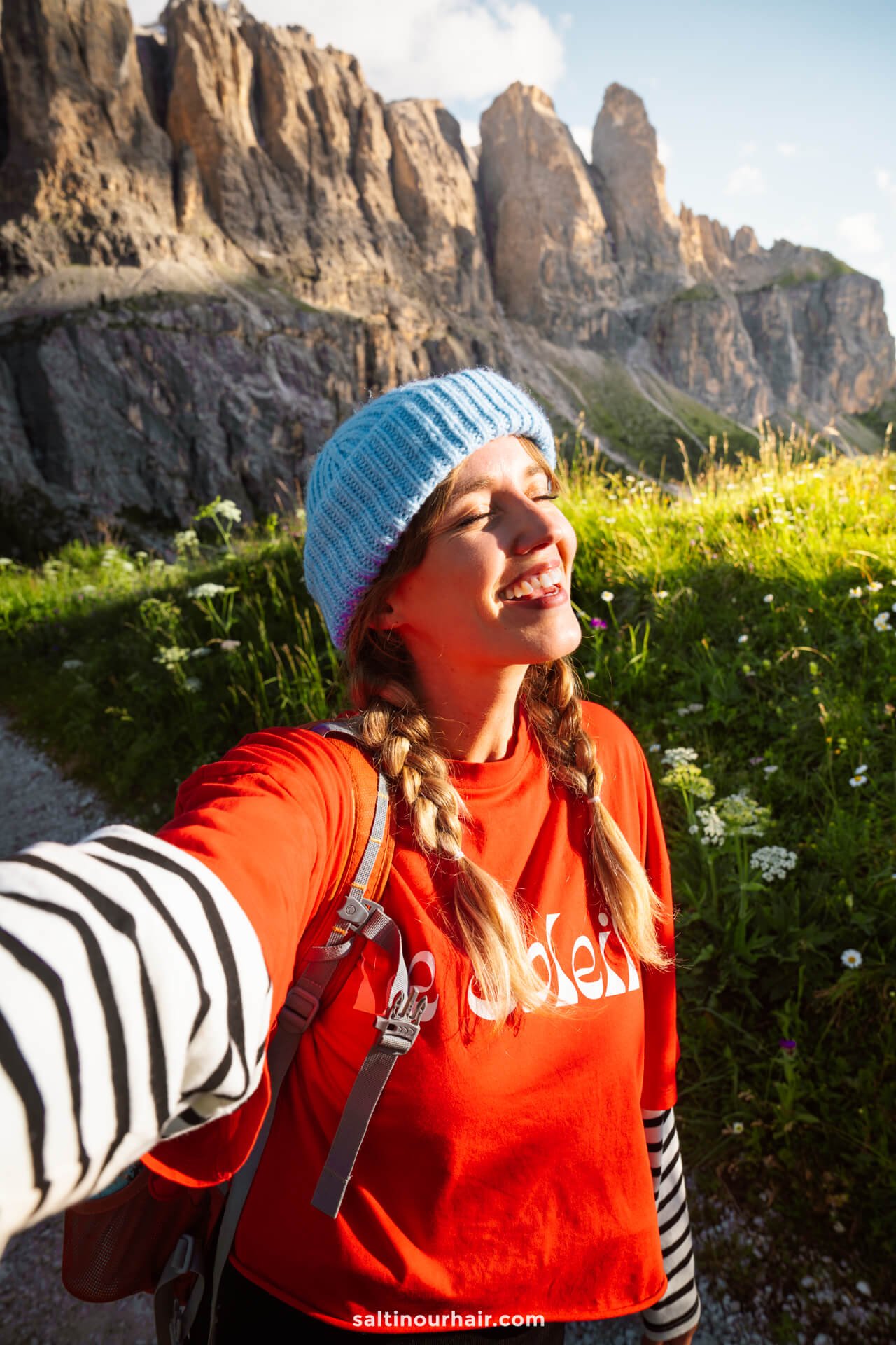 Woman wearing a blue knit beanie and red shirt smiles while taking a selfie outdoors with rocky mountains and wildflowers in the background, capturing the Gardena Pass. One of the unforgettable things to do in the Dolomites.