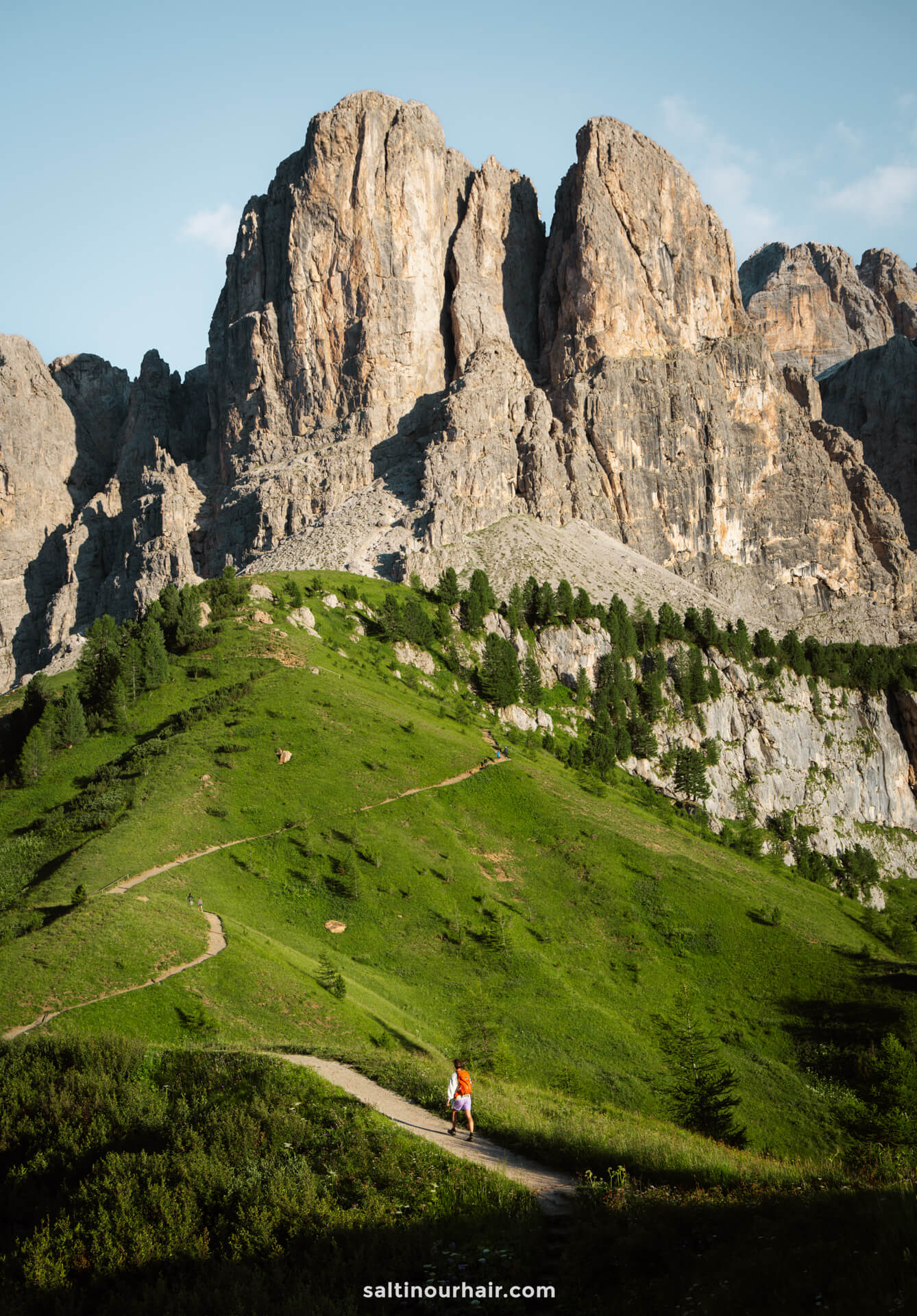 A man with a backpack walks along a winding trail on a green hillside, with steep rocky mountains of the Gardena Pass rising in the background—a perfect scene for those seeking things to do in the Dolomites under a clear sky.