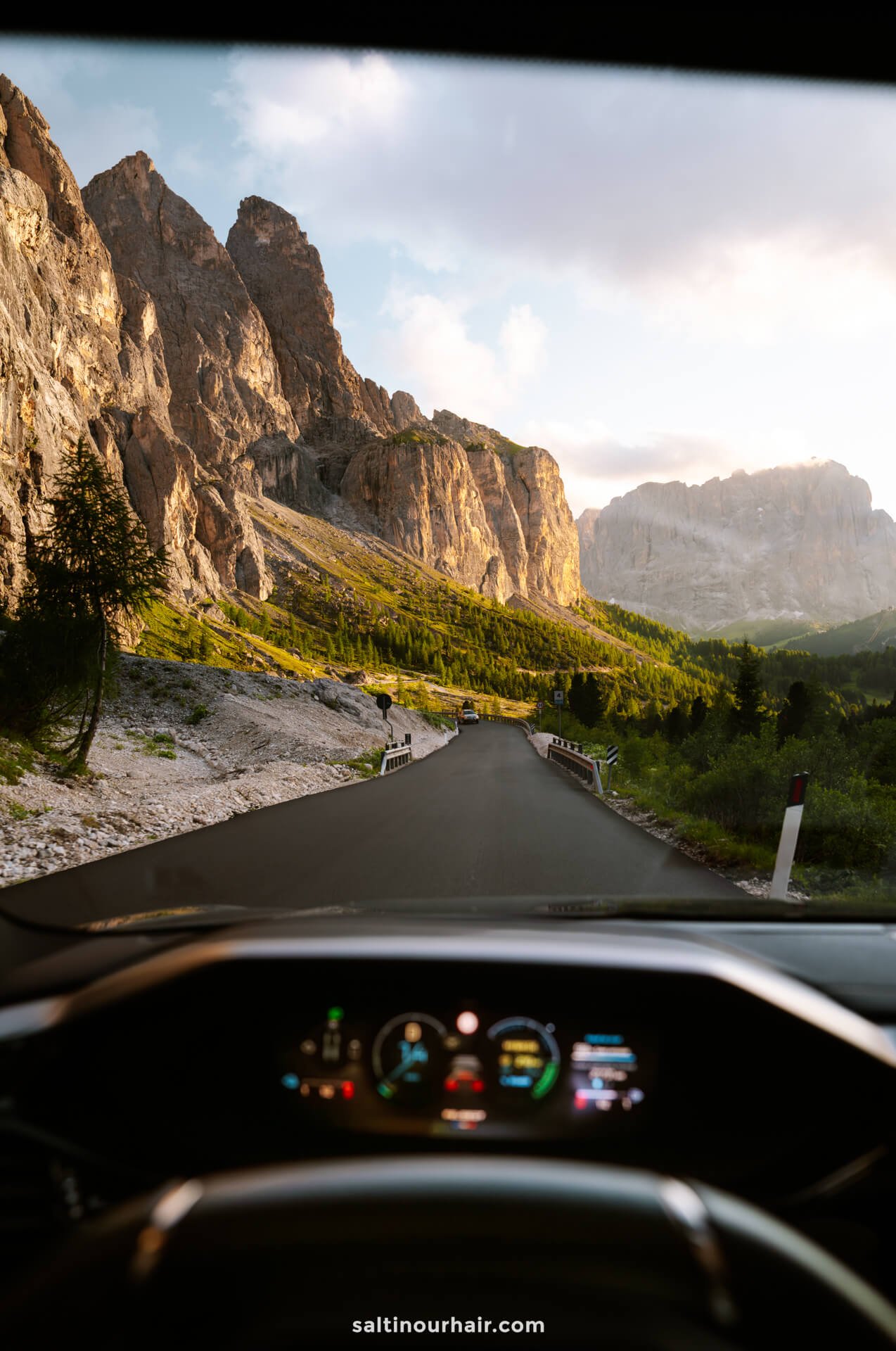 View from inside a car driving on a paved mountain road of the Gardena Pass in the Dolomites, with rocky cliffs and green trees on both sides under a partly cloudy sky