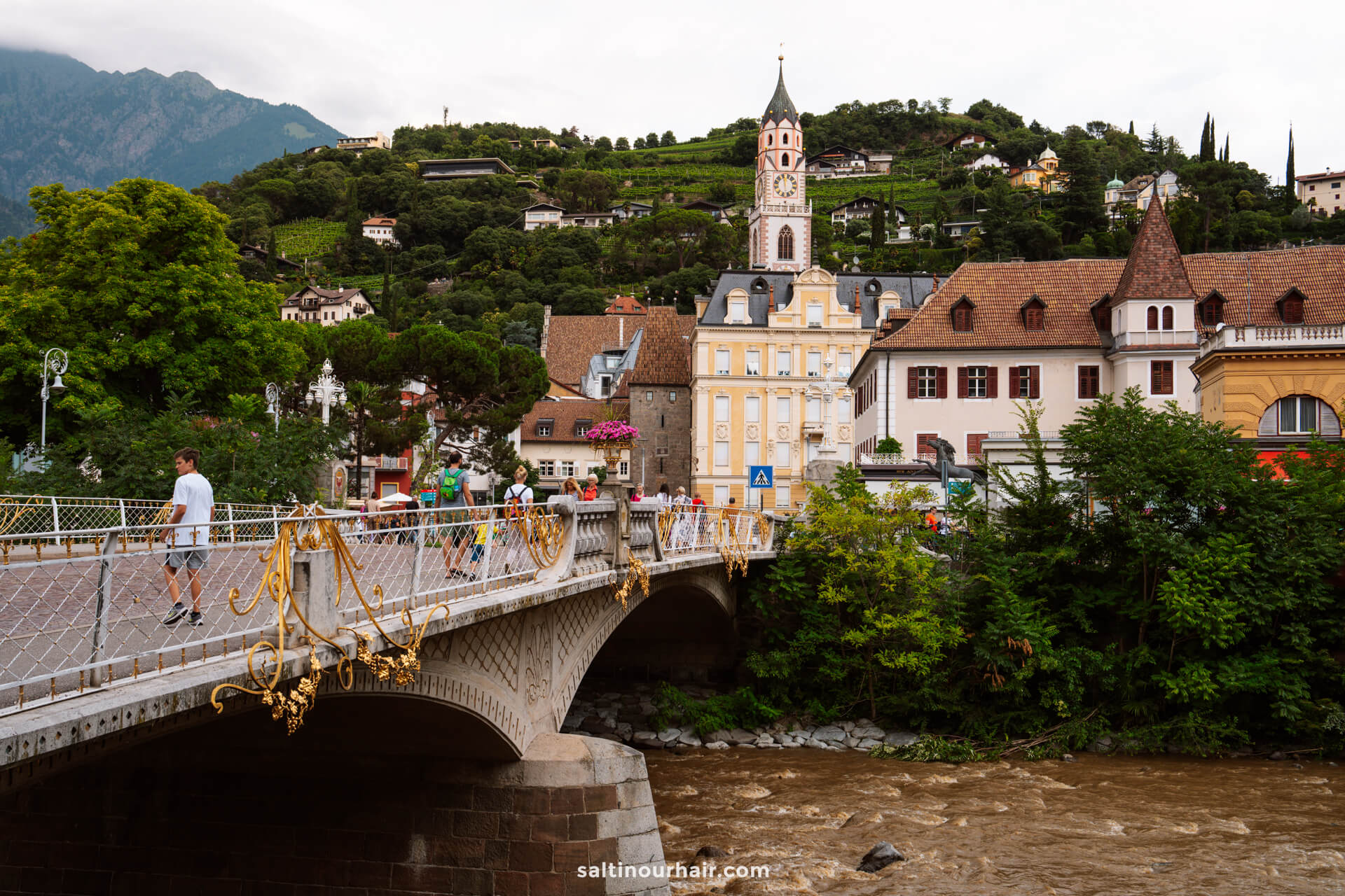 A pedestrian bridge with ornate railings crosses a brown river, leading to historic buildings and a clock tower set against a green, hilly backdrop—an inviting scene among the many things to do in the Dolomites.