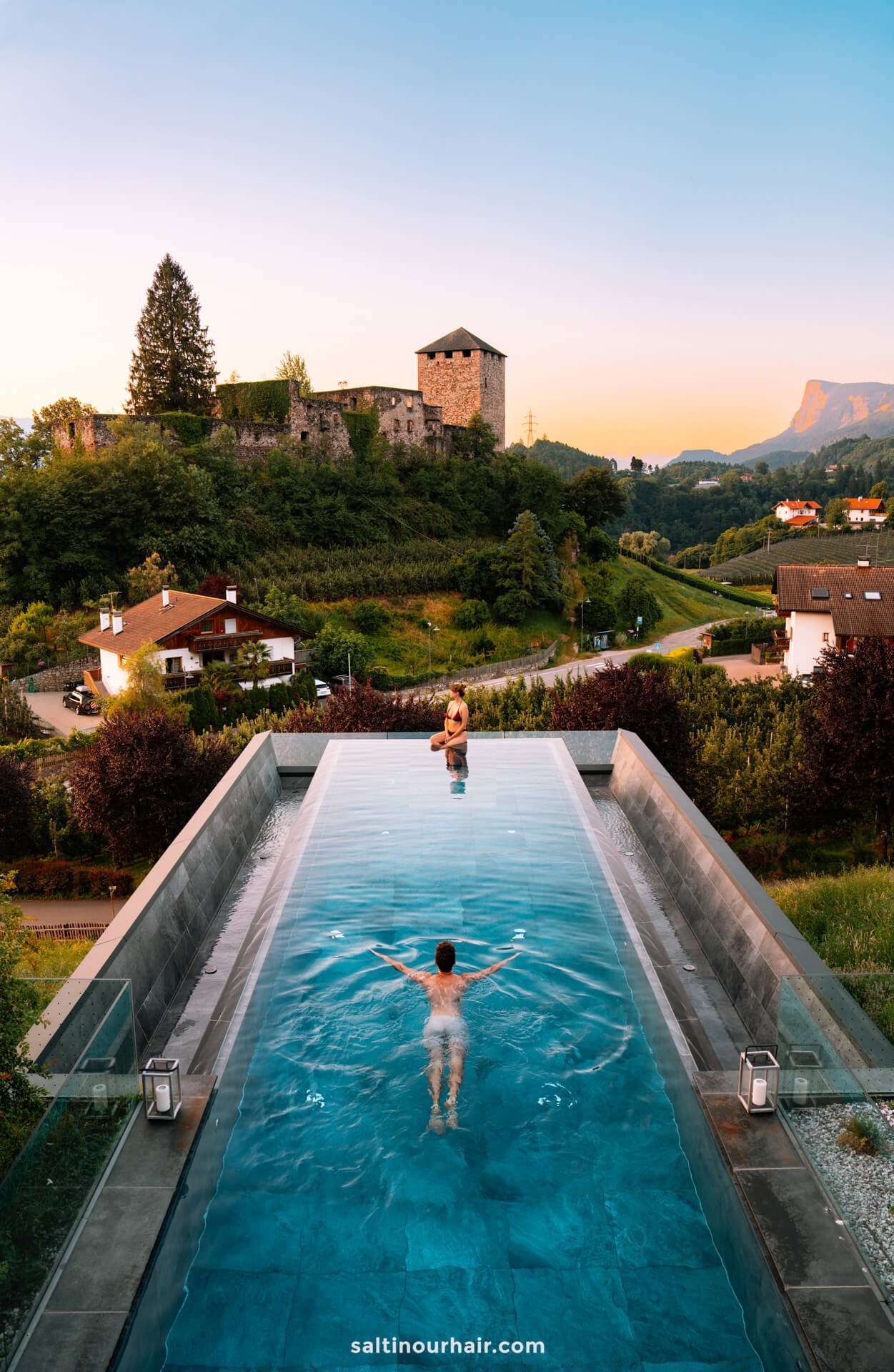Two people relax in a rectangular outdoor pool at Hotel der Waldhof overlooking a scenic Dolomite landscape of houses, greenery, and a stone building on a sunset-lit hill