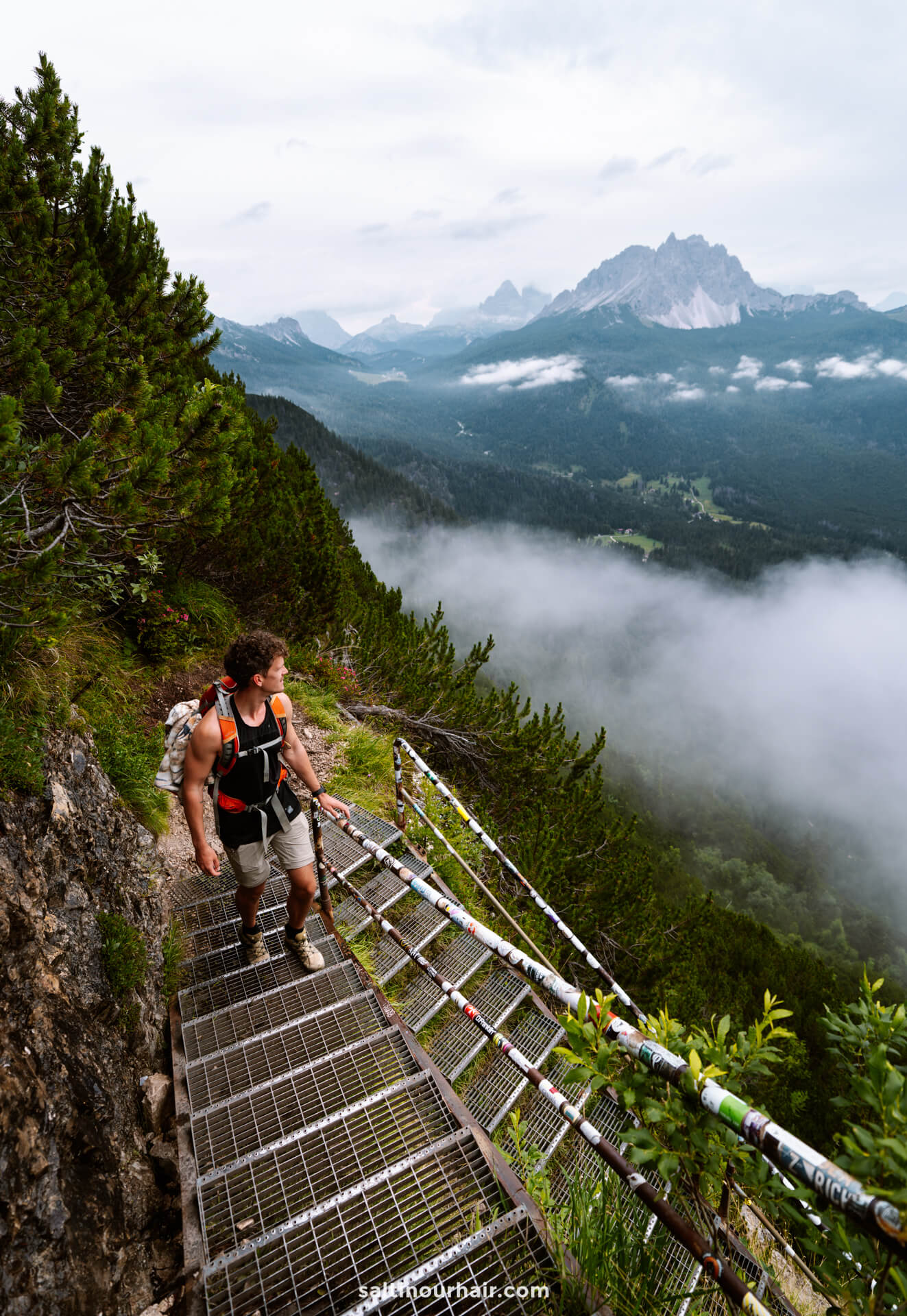 A hiker with a backpack climbs a metal staircase on a mountain trail to lago di sorapis in the Dolomites, surrounded by trees, mist, and distant peaks—a stunning adventure and one of the top things to do in the Dolomites italy