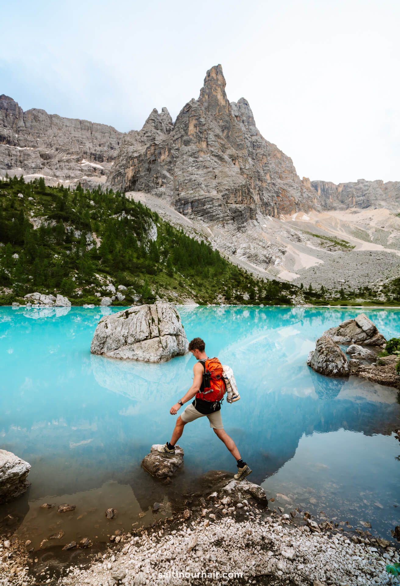 A man with a red backpack steps on rocks by a turquoise mountain lake called lago di sorapis, surrounded by steep rocky peaks and green trees—one of the unforgettable things to do in the Dolomites italy