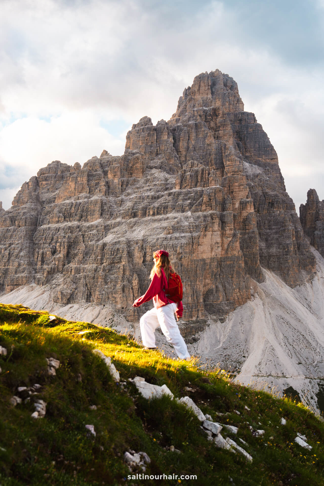 A woman wearing a red jacket and white pants hikes up a grassy slope with a large rocky mountain in the background—Cadini di Misurina is one of the unforgettable things to do in the Dolomites.