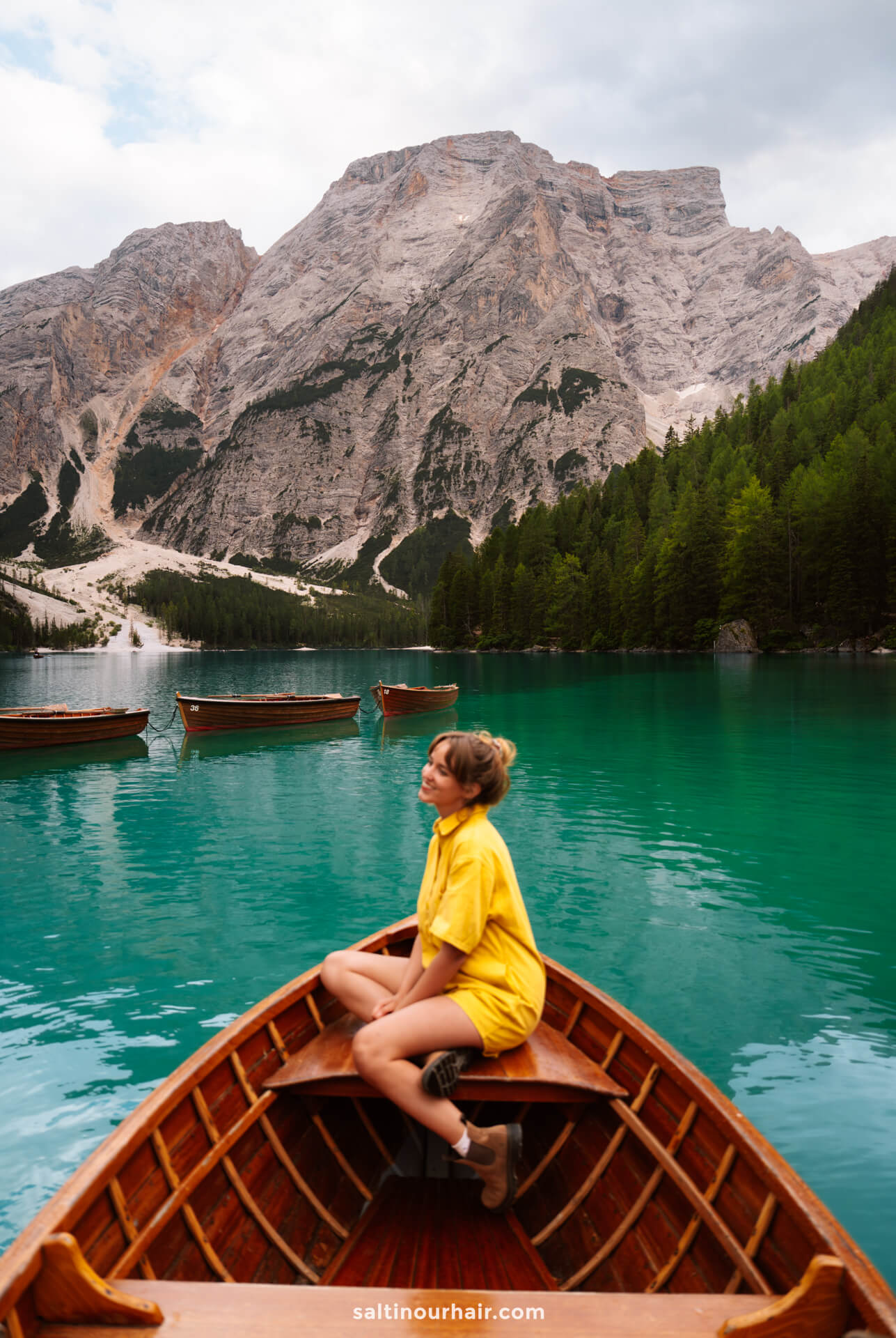 A woman in a yellow outfit sits at the front of a wooden boat on a turquoise lake called Lago di Braies, surrounded by mountains and pine trees—one of the unforgettable things to do in the Dolomites italy