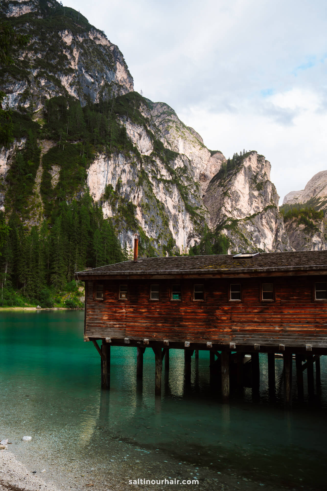 A wooden boathouse on stilts over a turquoise lake called Lago di Braies, surrounded by forested mountains and rocky cliffs under a partly cloudy sky—one of the most picturesque things to do in the Dolomites italy