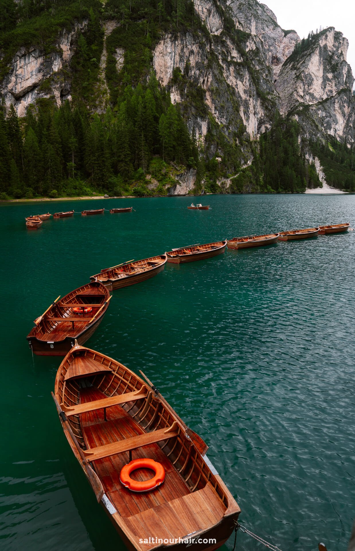 Wooden rowboats are lined up in a curved formation on the turquoise lake of lago di braies, framed by pine forest and rocky mountains under an overcast sky—an iconic scene among the unforgettable things to do in the Dolomites.