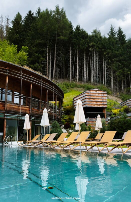 Outdoor pool with yellow lounge chairs and white umbrellas, modern wooden buildings, and a backdrop of dense forest under a cloudy sky—an idyllic spot for relaxing after exploring the top things to do in the Dolomites.
