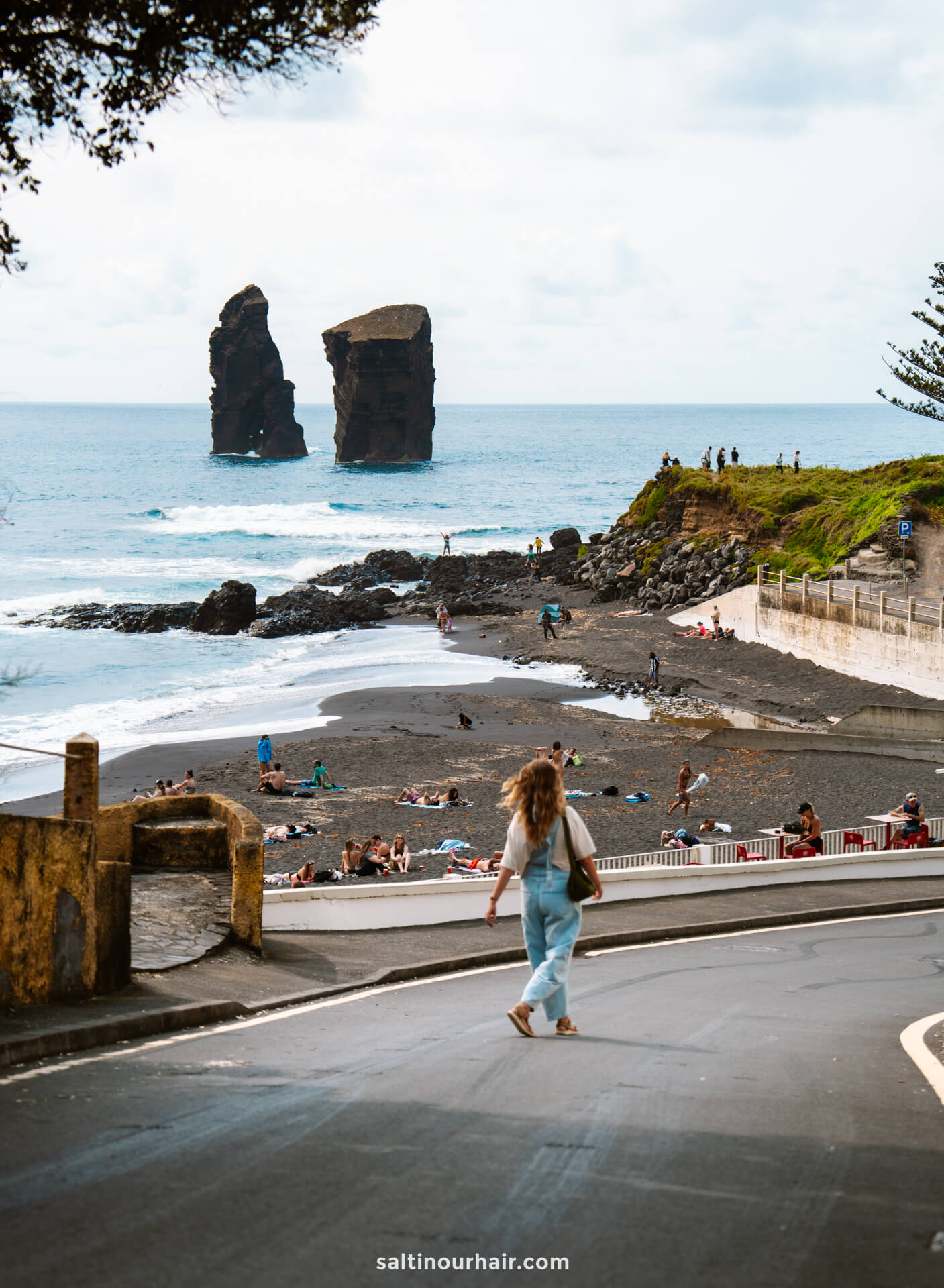 A woman crosses a road toward a black sand beach—an essential stop on any São Miguel itinerary—with people sunbathing and swimming; two large sea stacks rise from the ocean in the background.
