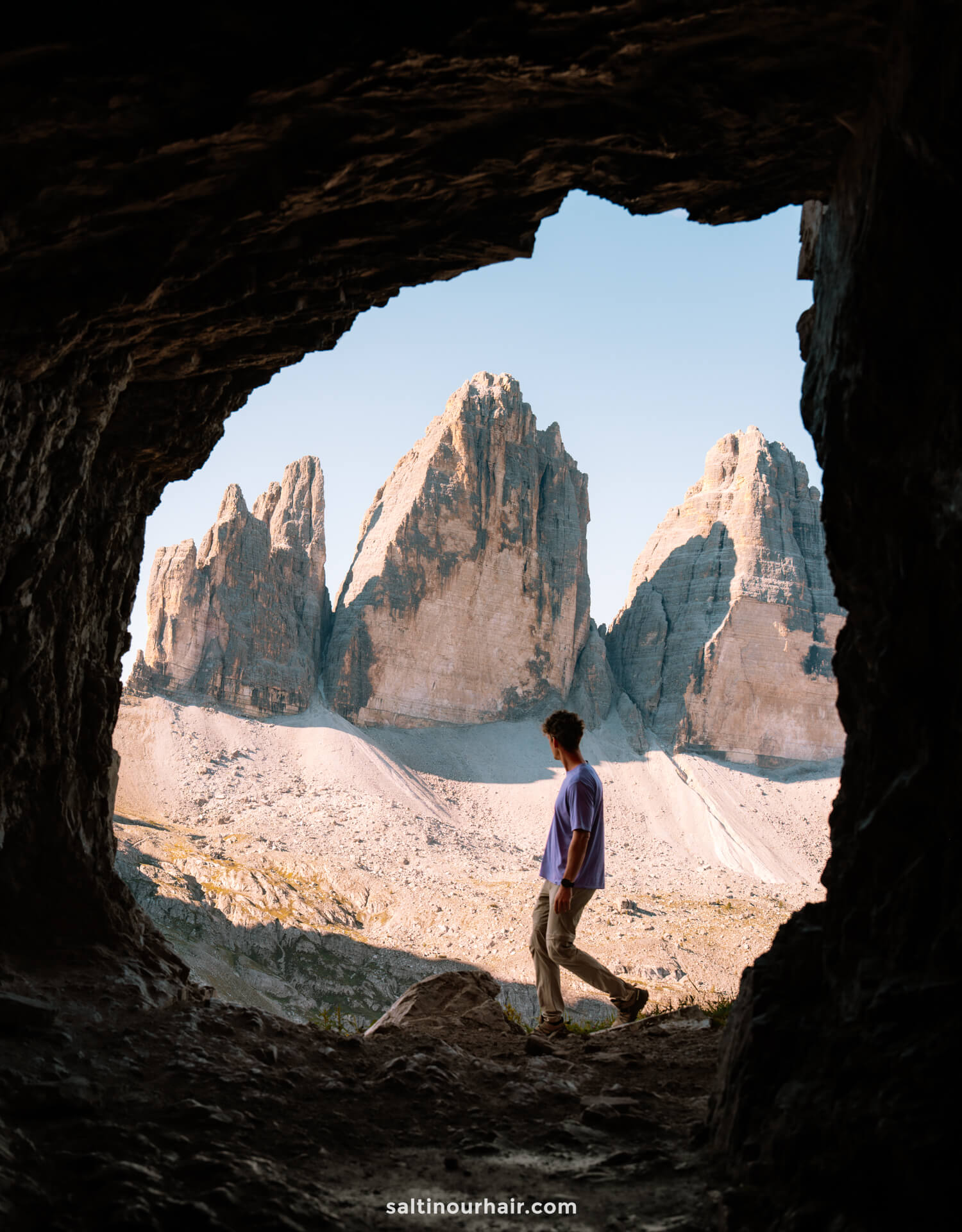 A person stands at the entrance of a cave, looking out at the iconic Tre Cime peaks rising sharply under a clear sky.