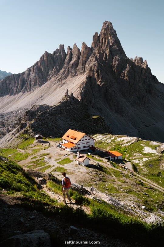 A person hikes uphill on a trail toward a group of alpine buildings, with the iconic Tre Cime’s jagged mountain peaks rising in the background under a clear sky.