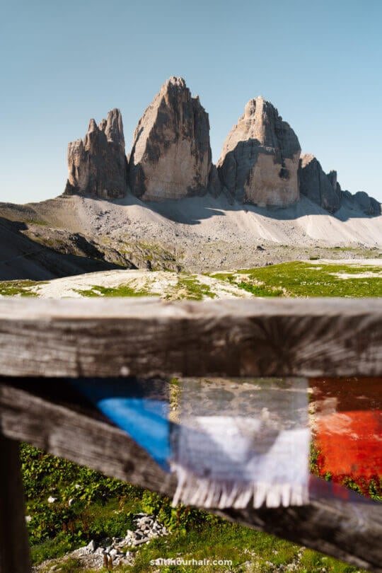 A wooden fence with faded fabric in the foreground frames the iconic Tre Cime peaks and green grass under a clear blue sky in the background.