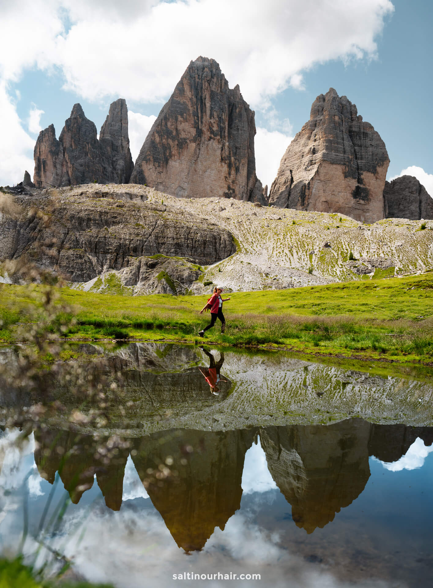 Person in a red jacket walks by a small pond reflecting the Tre Cime peaks and partly cloudy sky, set within a grassy, rocky landscape.