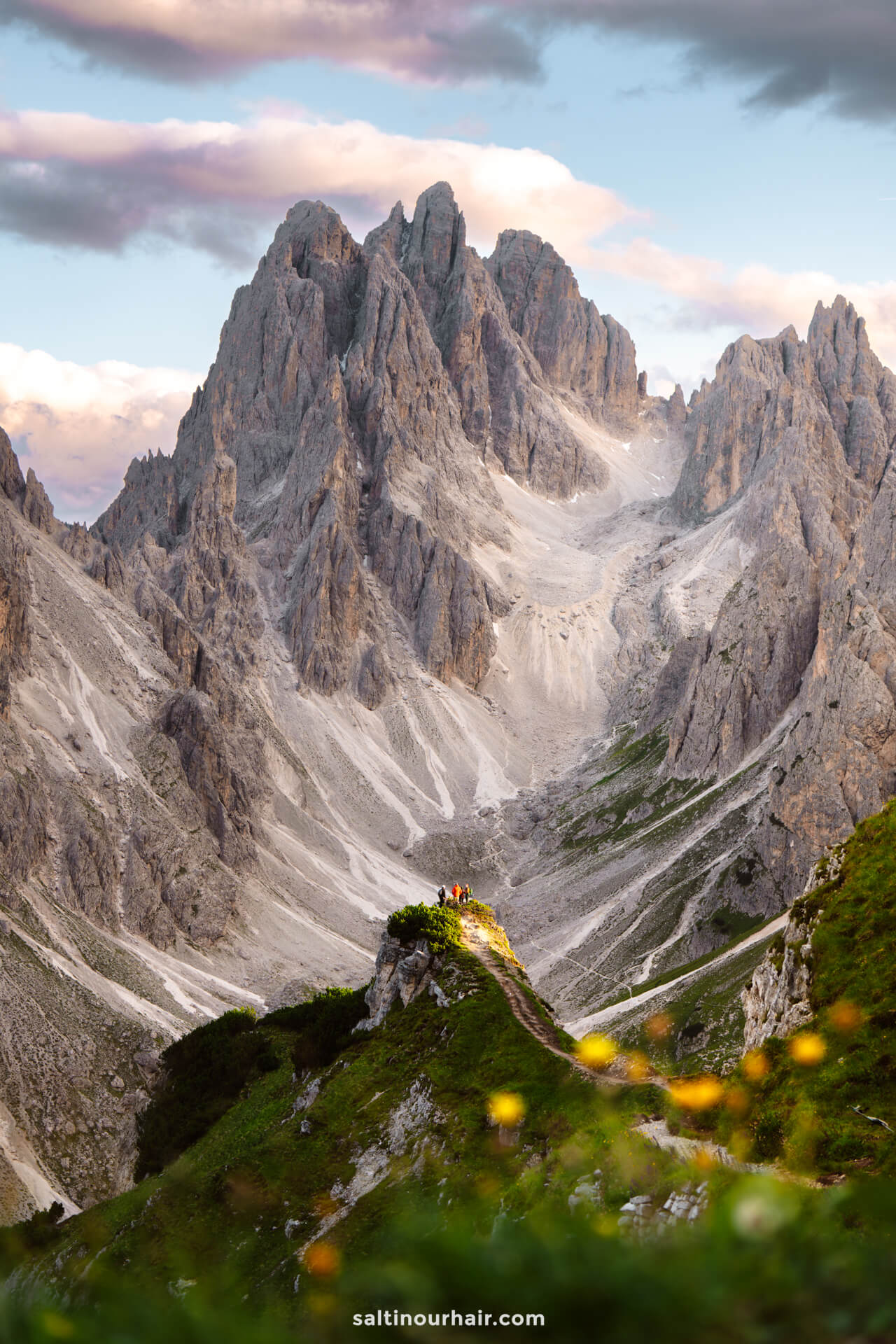 A group of people stands on a grassy ridge overlooking the dramatic, rocky peaks of Tre Cime, set against a partly cloudy sky.