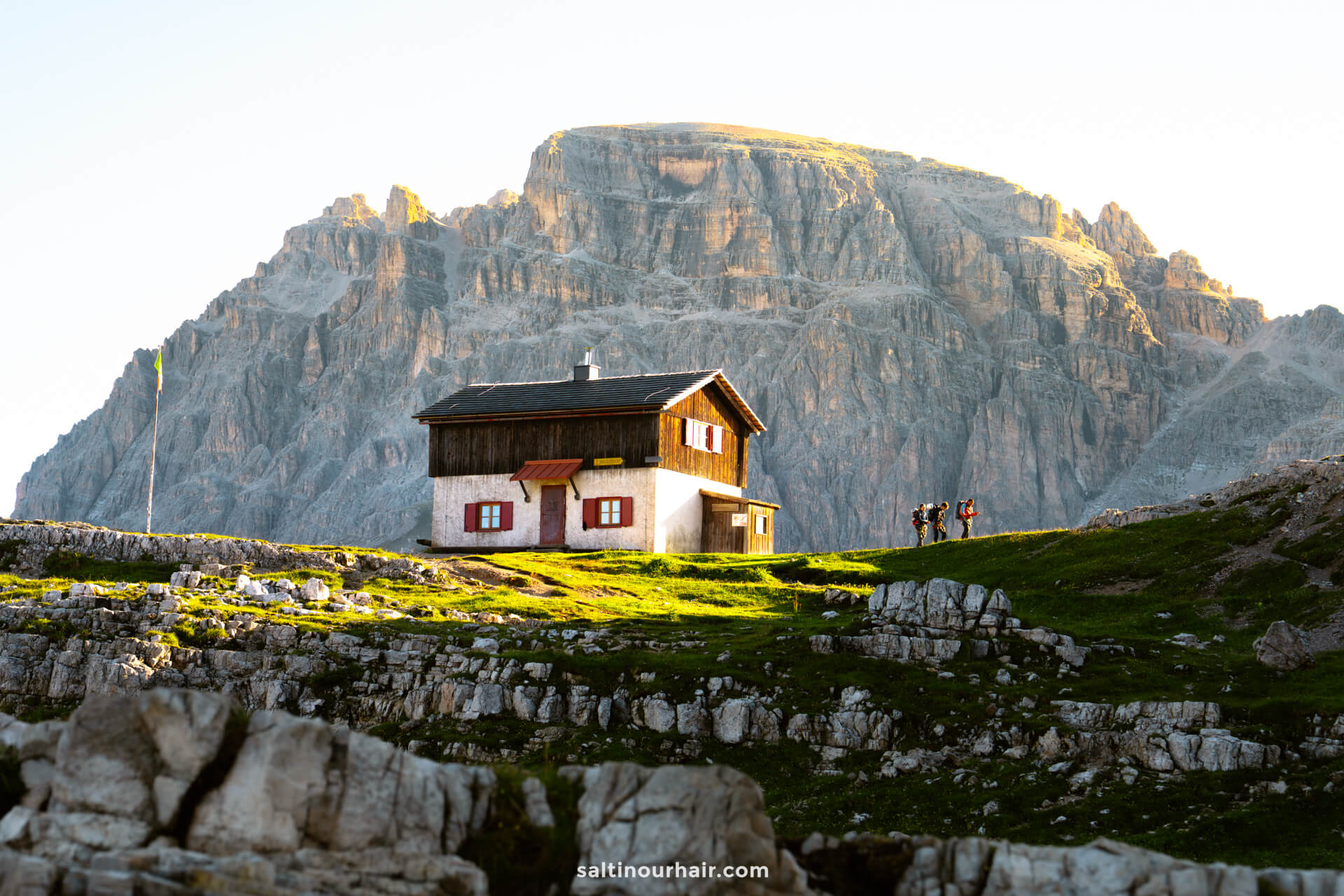 A small house sits on a grassy hill with large rocky mountains, including the iconic Tre Cime, in the background and a group of hikers standing nearby.