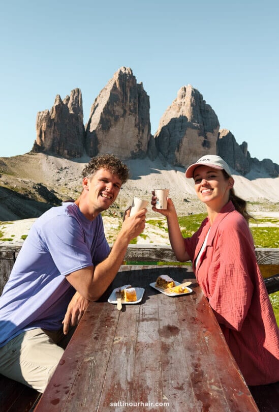 Two people sit at a wooden picnic table outdoors, smiling and toasting with cups, with the iconic Tre Cime mountain peaks visible in the background. Food is spread on the table, creating a perfect alpine picnic scene.