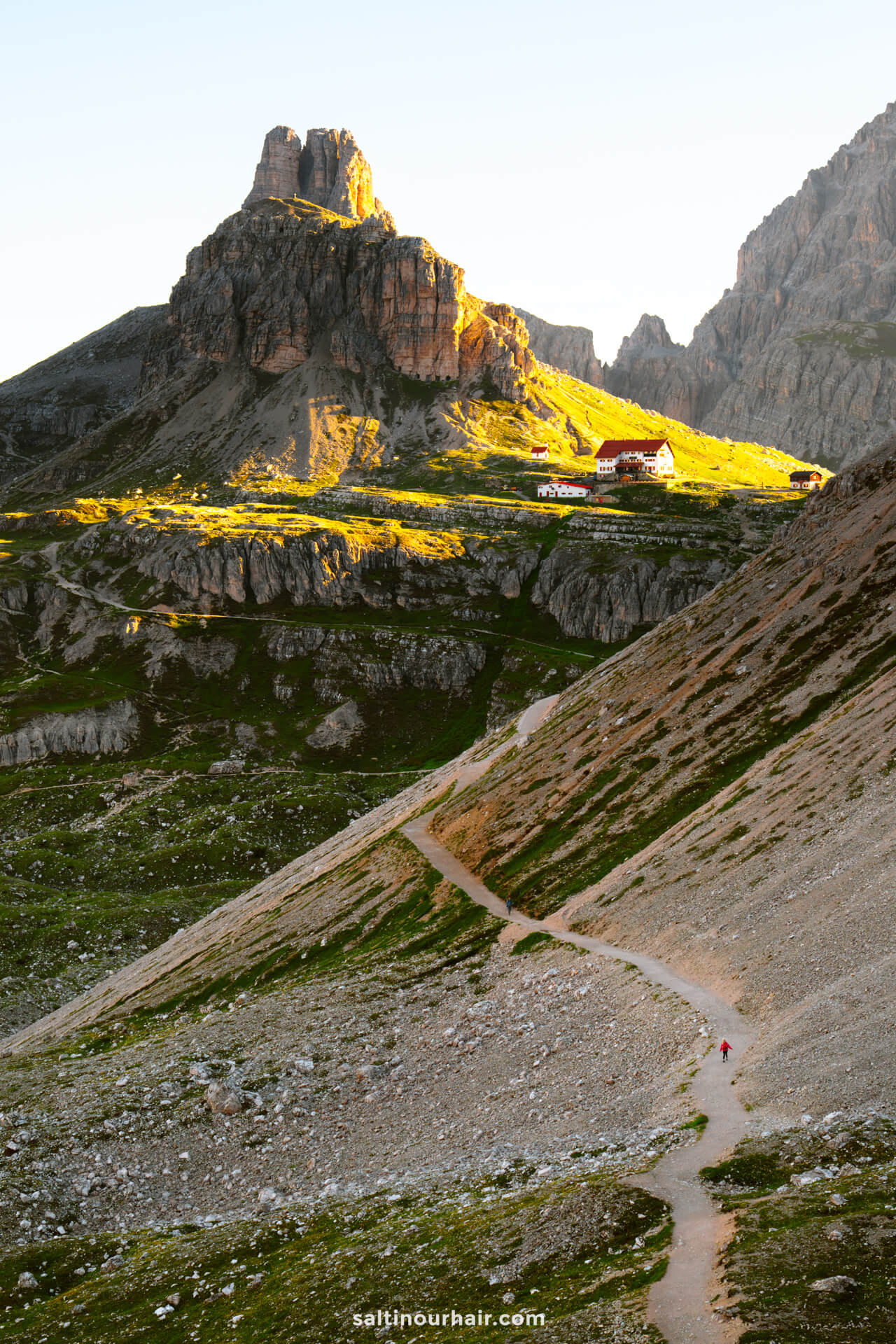 A winding mountain trail leads to a sunlit building nestled below the rocky peaks of Tre Cime, all beneath a clear sky.