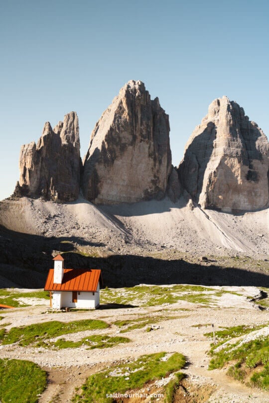 A small white chapel with a red roof stands in front of the iconic Tre Cime peaks, three tall rocky mountains rising under a clear blue sky.