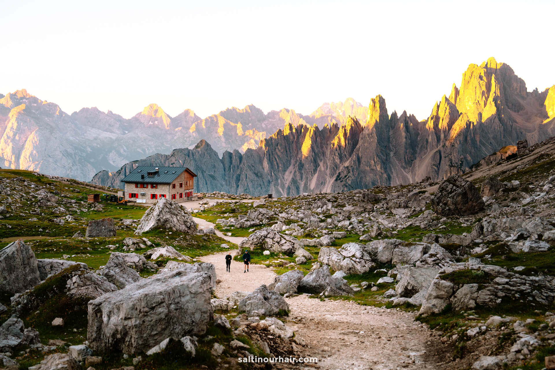Two people walk on a rocky trail toward a mountain lodge, surrounded by the rugged peaks of Tre Cime and illuminated by warm sunlight in the background.