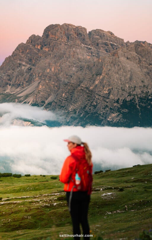 A person in a red jacket stands on a grassy field with Tre Cime’s iconic peaks and low clouds in the background.