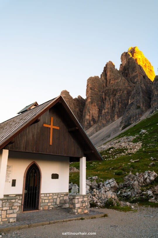 A small chapel with a wooden cross stands before the majestic Tre Cime, its silhouette illuminated by the golden light of sunset against the rocky mountain peaks.
