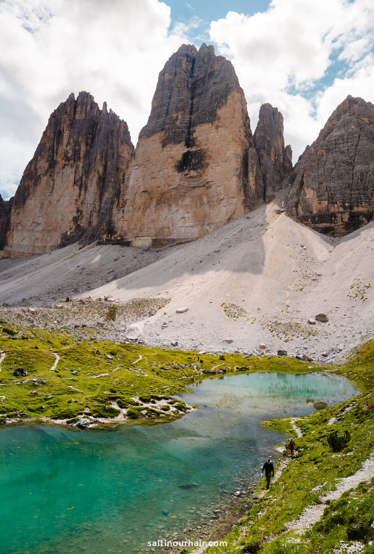 A clear turquoise lake sits at the base of tall, rugged mountain peaks, including the iconic Tre Cime, with two people walking along the grassy edge under a partly cloudy sky.