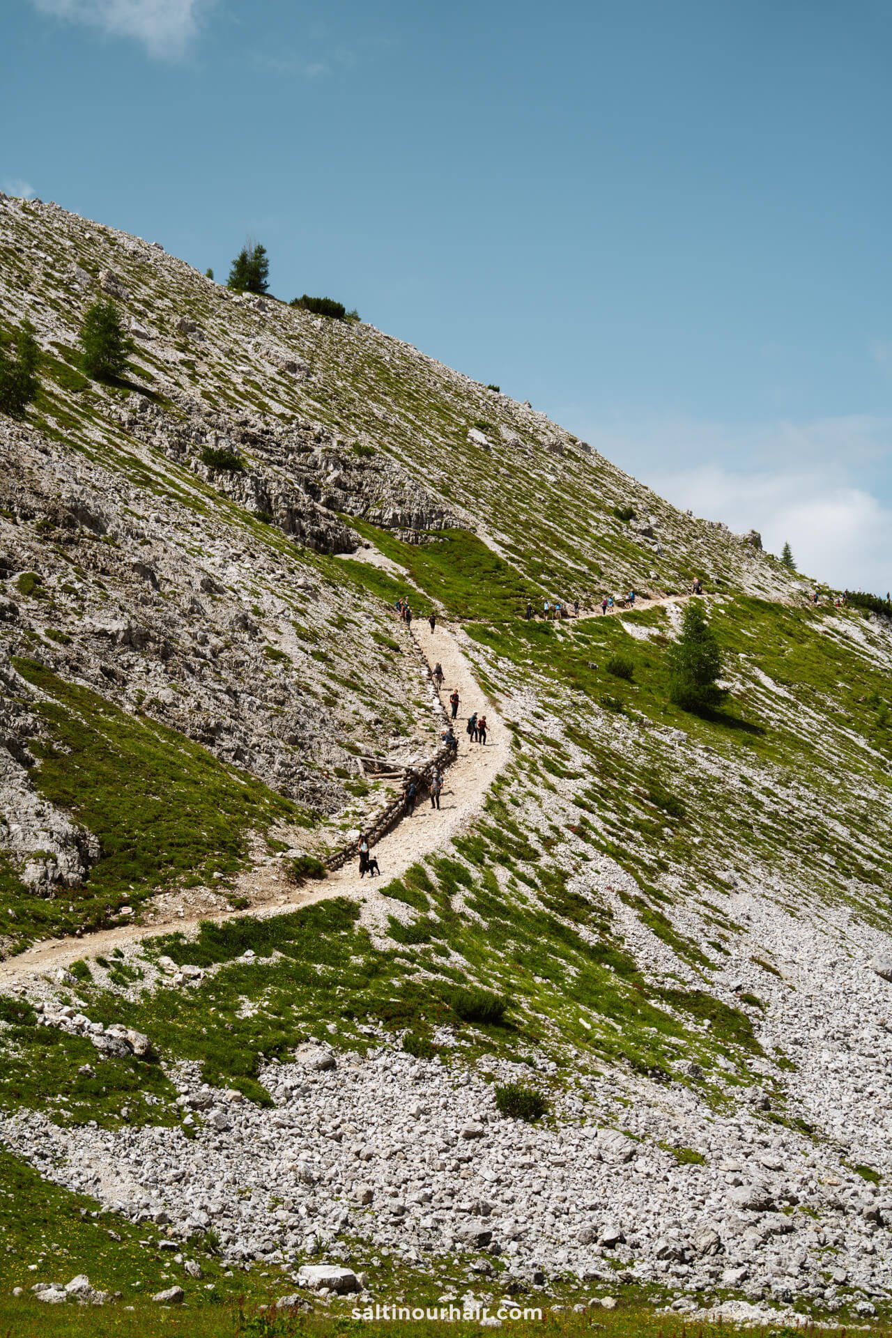 A group of hikers walk along a winding trail on a rocky, grassy hillside beneath the iconic peaks of Tre Cime and a partly cloudy sky.
