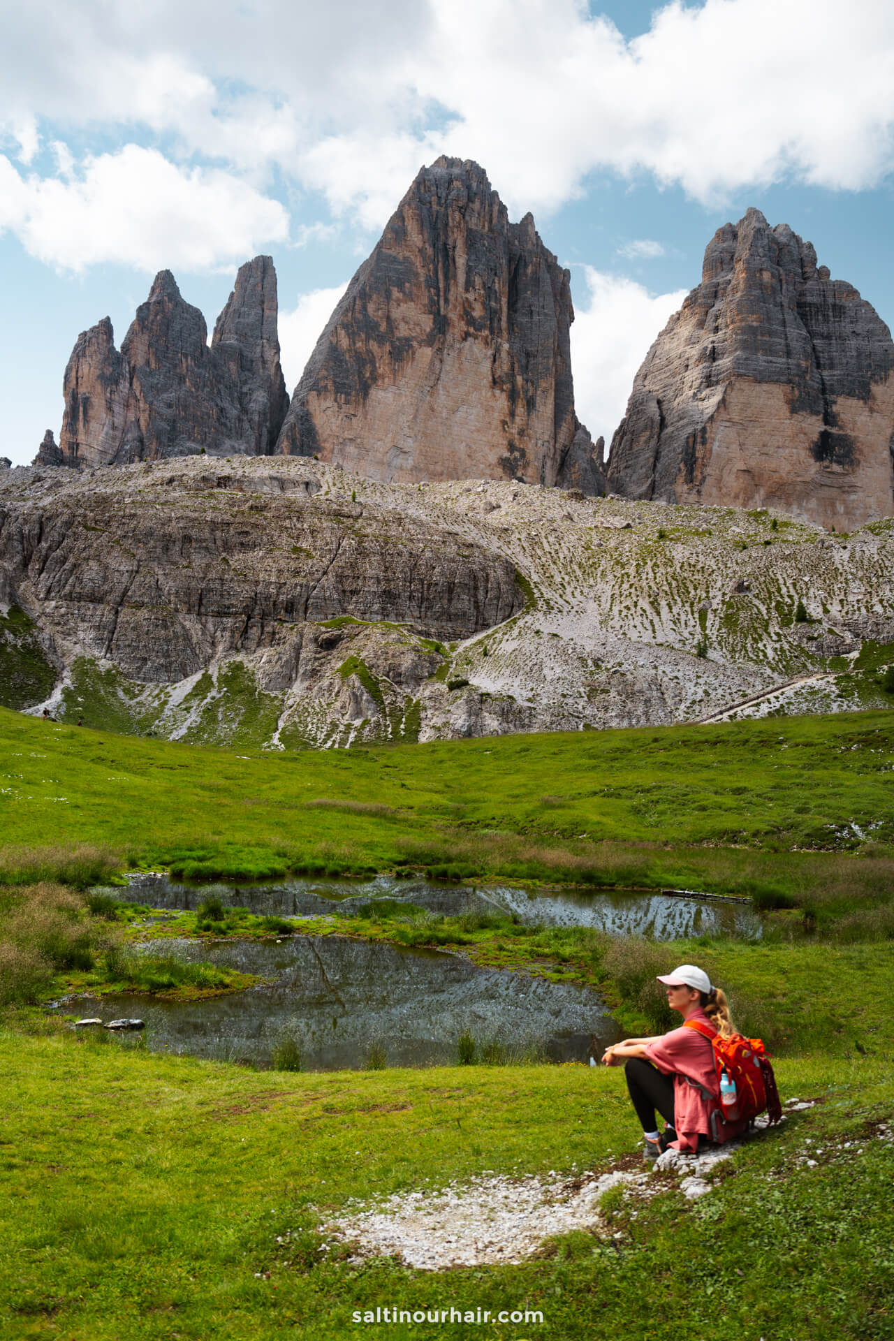 A person with a red backpack sits on grass near a small pond, with the iconic Tre Cime’s rocky peaks rising in the background under a partly cloudy sky.