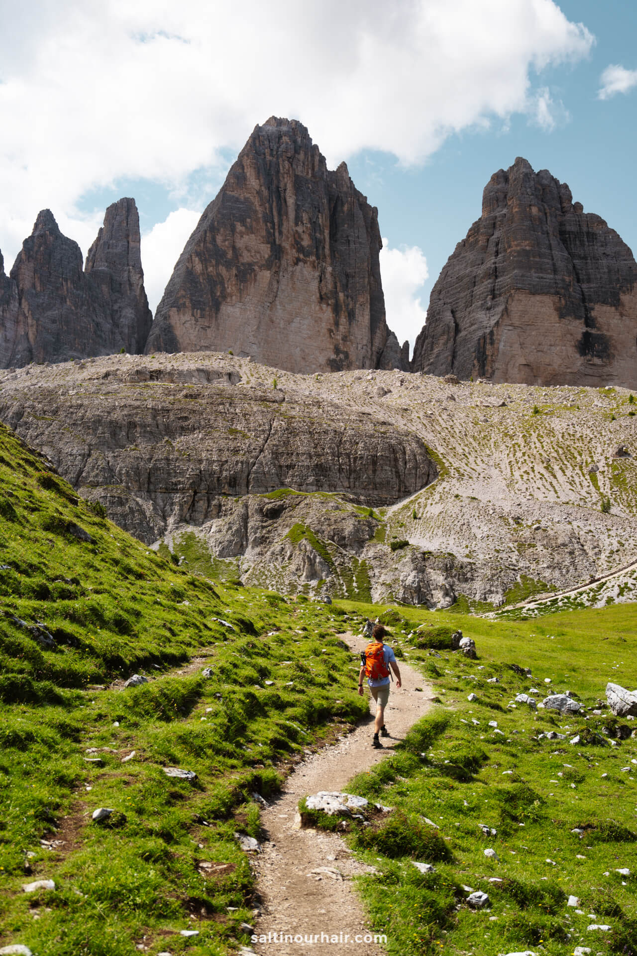A person with a backpack walks along a dirt path through grassy hills, with the iconic peaks of Tre Cime and a partly cloudy sky in the background.