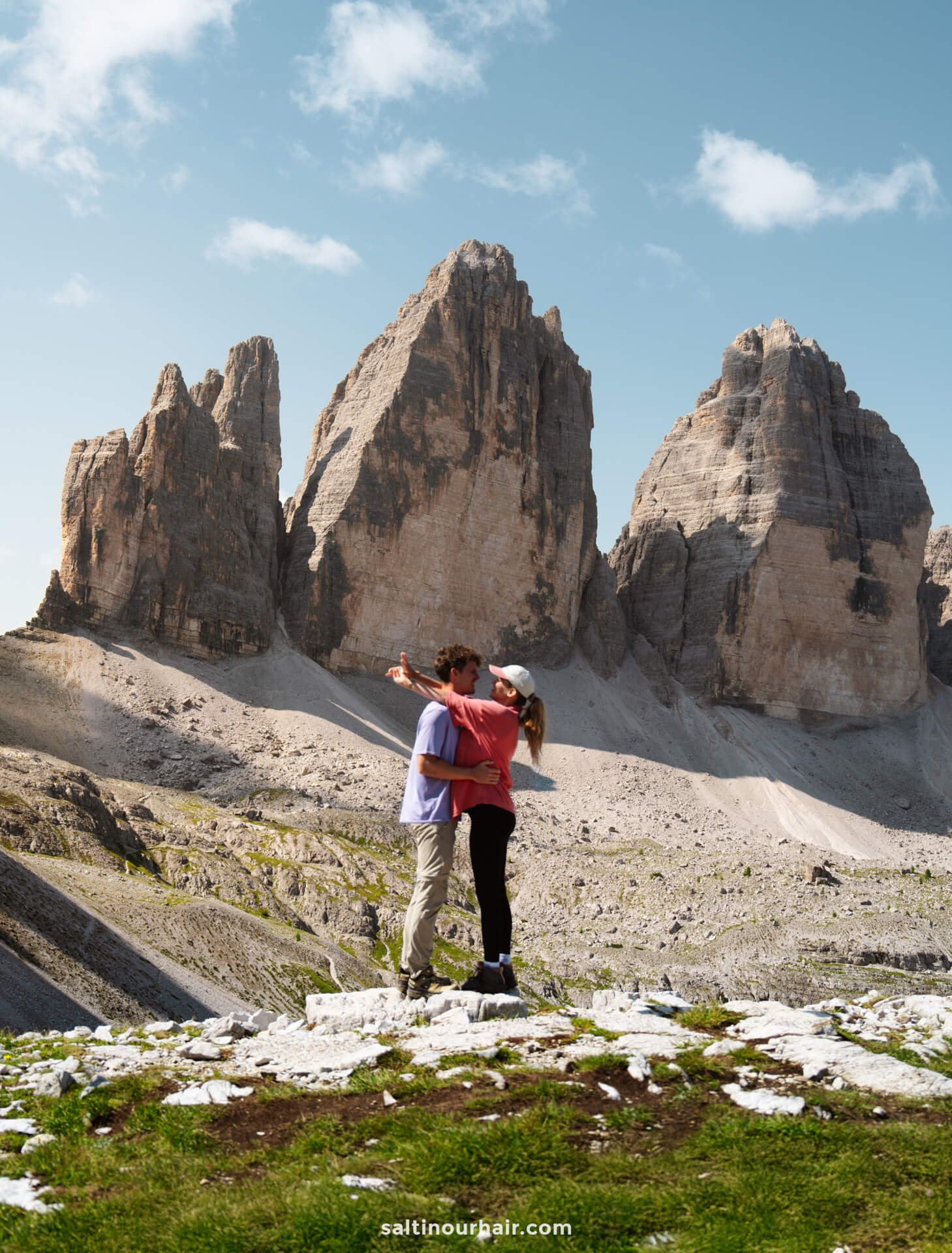 A couple stands on rocky terrain, with one person pointing towards the iconic Tre Cime peaks in the background beneath a partly cloudy sky.