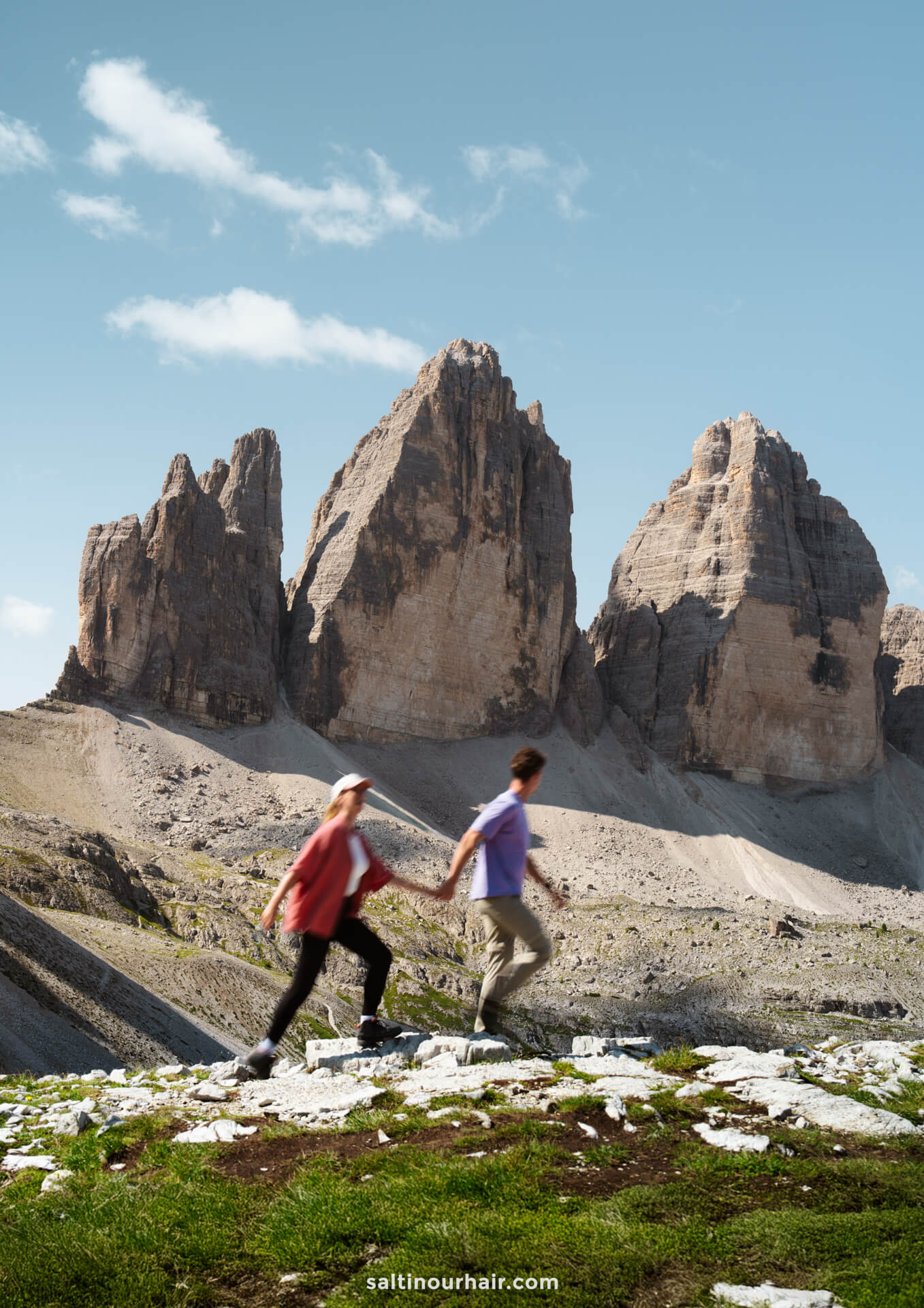 A couple walks in the foreground, framed by the majestic rocky peaks of Tre Cime rising in the background beneath a clear sky.