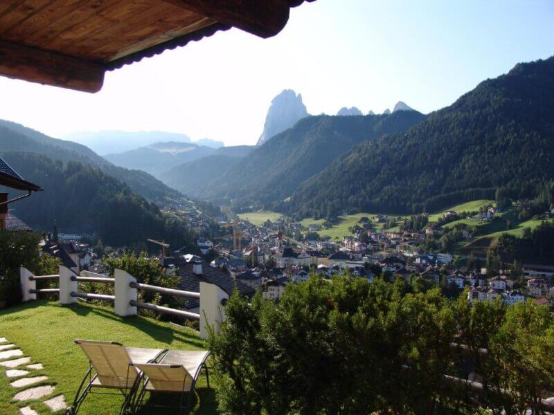 A scenic mountain valley view from a terrace with two lounge chairs, overlooking the town of Ortisei surrounded by green hills and the distant peaks of the Dolomites under a clear sky.
