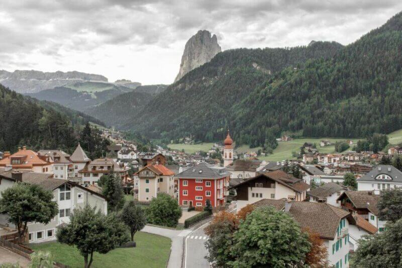 A mountain village in the Dolomites with colorful houses, a church with a red dome, green hills, and a rocky peak in the background under a cloudy sky.