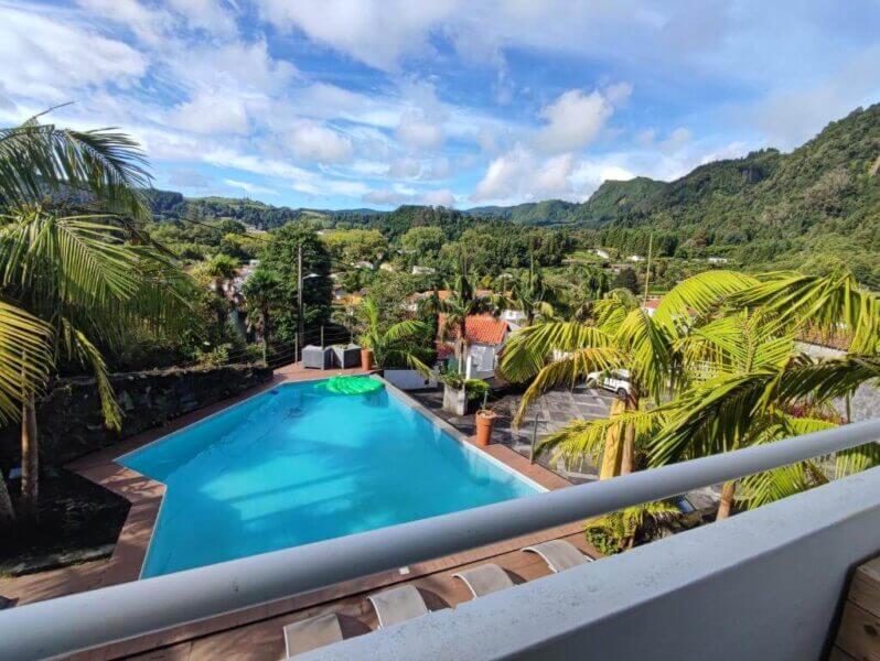 Rectangular outdoor swimming pool surrounded by palm trees, with a scenic landscape of hills and houses in the background under a partly cloudy sky—enjoy this relaxing Vista do Vale Furnas.