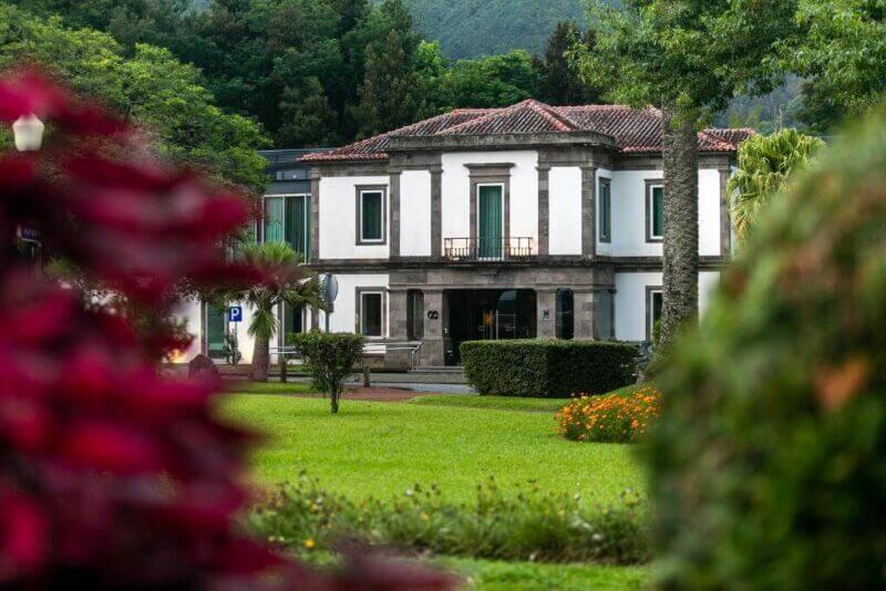 A two-story white building with green trim, part of Octant Furnas, stands amid a landscaped garden with grass, trees, flowers, and shrubs; forested hills are visible in the background.