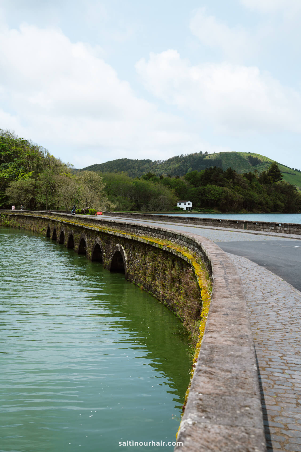 A curved stone bridge with multiple arches crosses over green water in Sete Cidades, with a road and a parked white vehicle in the background