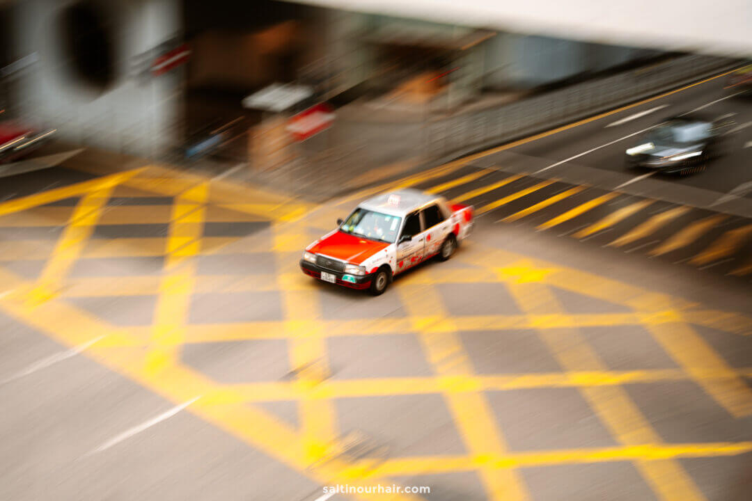 A red and white taxi speeds through a yellow crosshatched intersection in an urban area&mdash;an iconic scene to spot on your Hong Kong itinerary