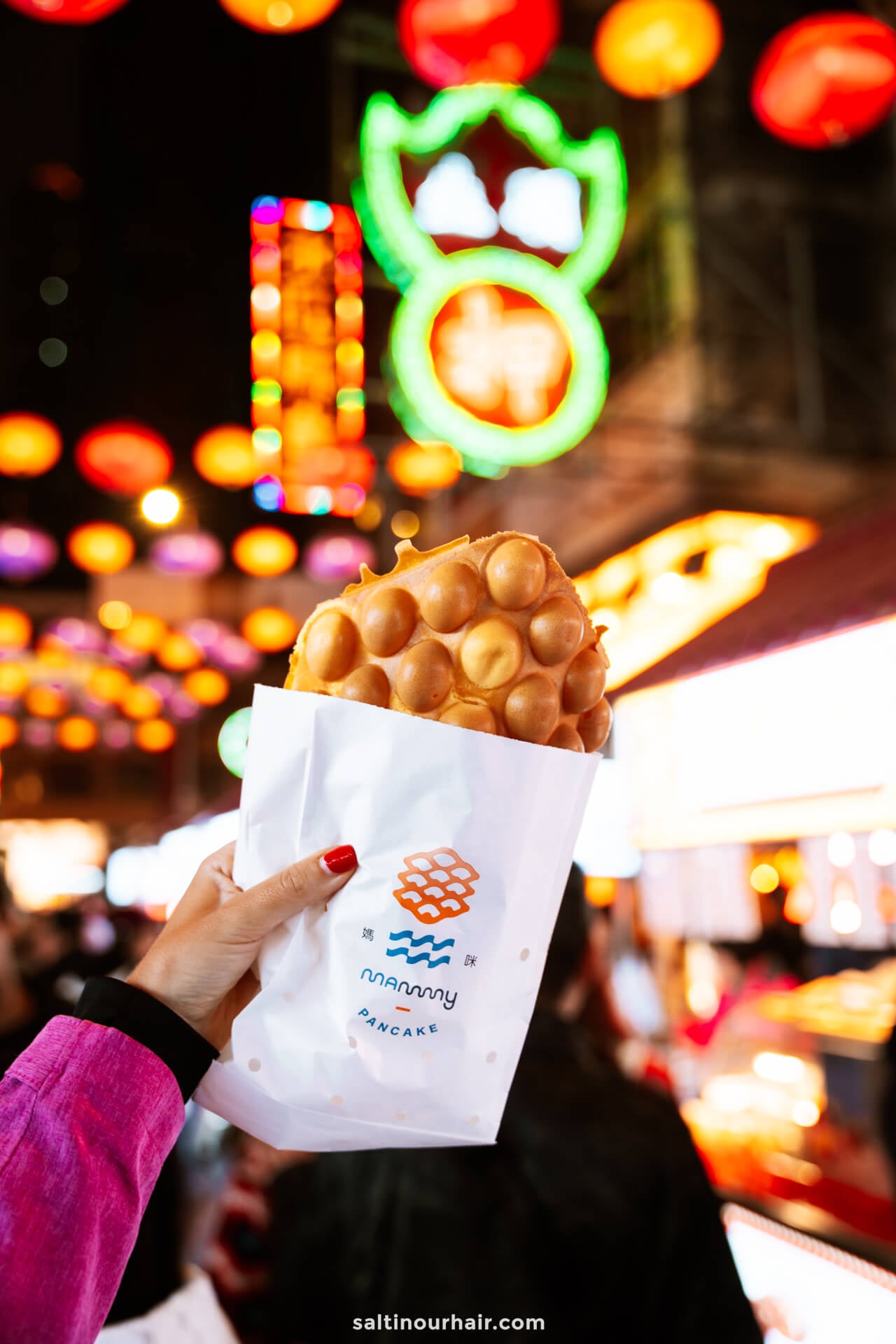 A hand holds a bubble waffle in a white paper sleeve, with colorful neon lights and lanterns glowing in the background&mdash;an iconic taste  of Hong Kong