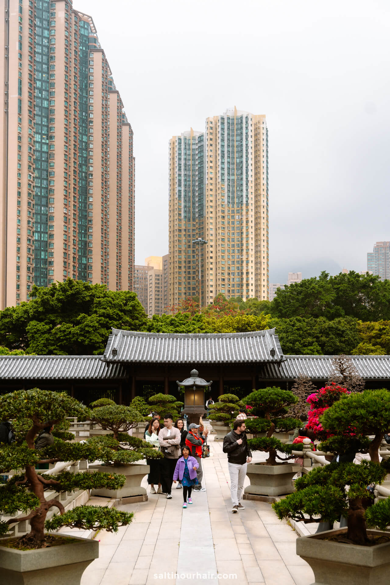 People walk through a landscaped garden with trimmed trees at Chi Linn Nunnery; tall modern apartment buildings rise in the background under a cloudy sky&mdash;a serene stop to include on your Hong Kong itinerary.