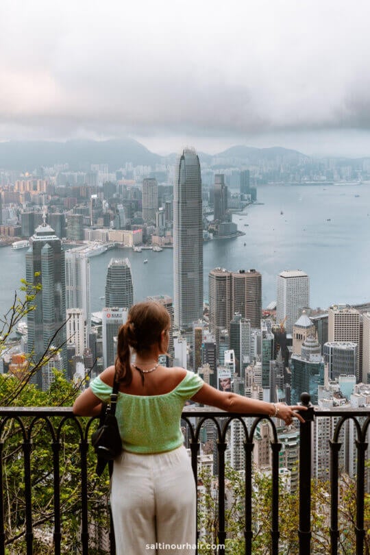 A woman stands at a railing overlooking the Hong Kong skyline and Victoria Harbour on a cloudy day, capturing a perfect moment for any hong kong itinerary.