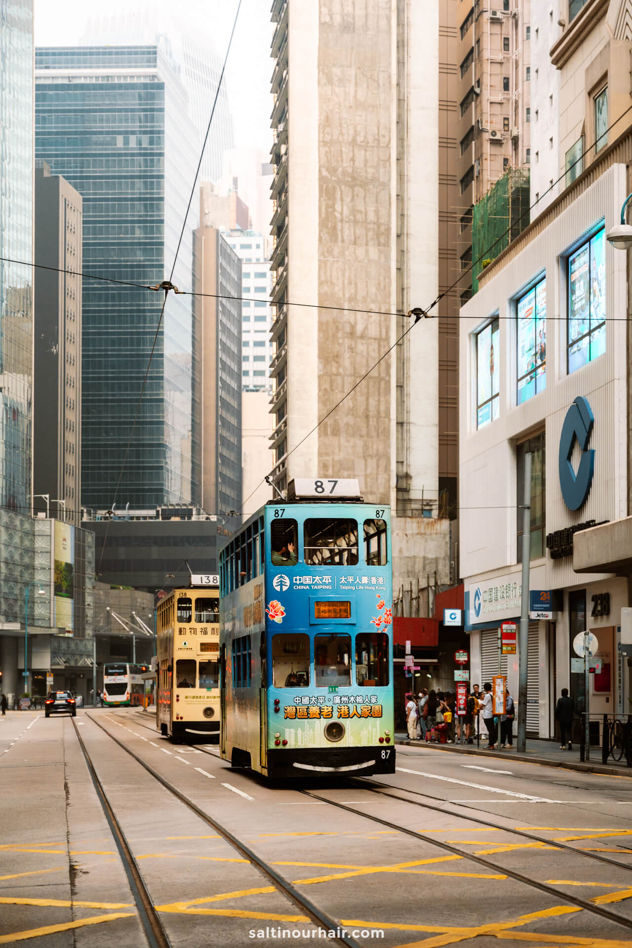 Double-decker trams travel along tracks on Central Hong Kong lined with tall buildings as pedestrians stroll along the bustling sidewalks.
