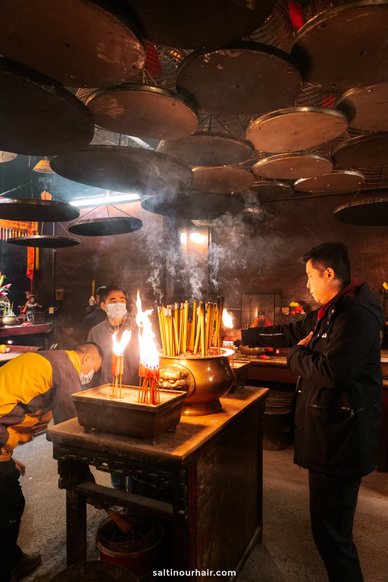 Three people stand near an incense altar inside Man Mo temple, with large incense coils hanging from the ceiling and smoke rising around them