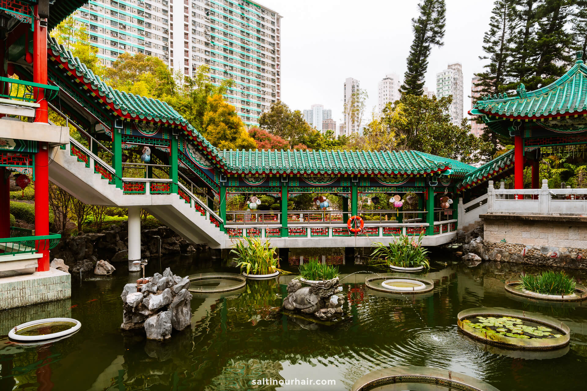 A traditional Chinese pavilion in Hong Kong with green tiled roofs sits over a pond with lily pads and rocks, surrounded by trees and modern high-rise buildings
