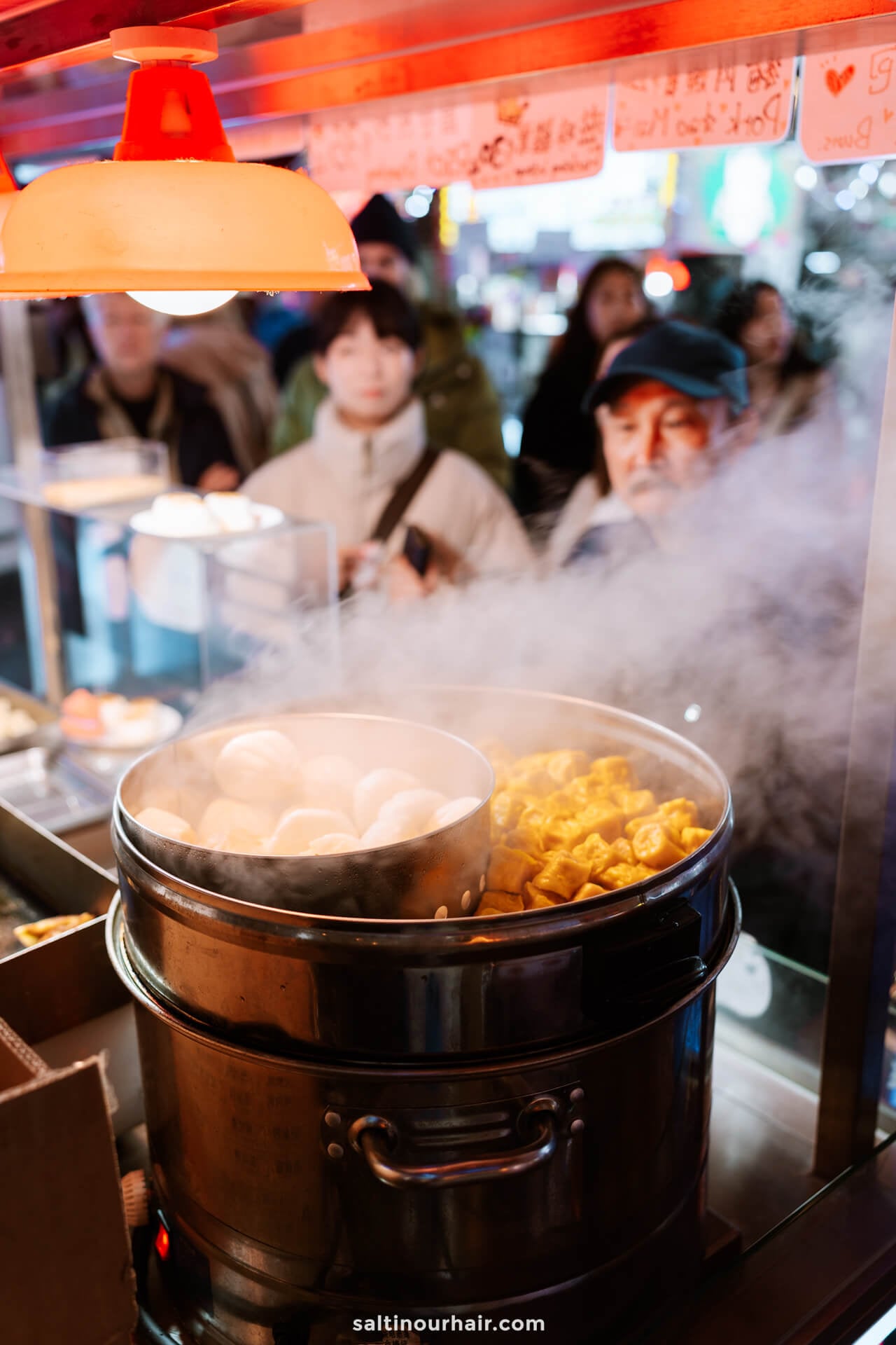 A steaming street food stall serves buns and dumplings in metal steamers as people wait in line