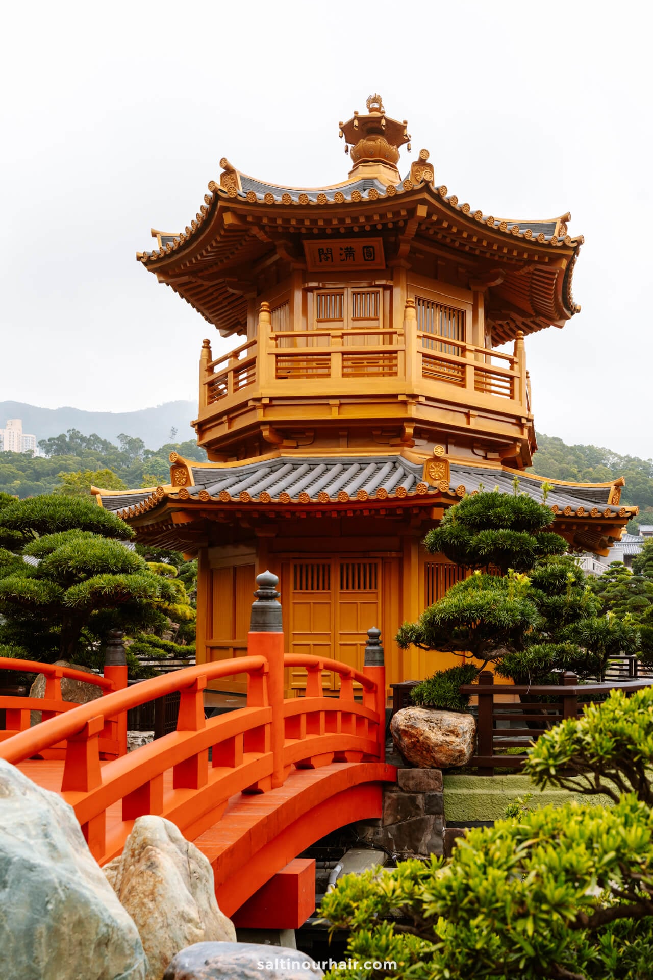 A traditional Chinese pavilion stands by a red arched bridge, surrounded by manicured trees, while city buildings and a misty mountain at Nan Lian Garden