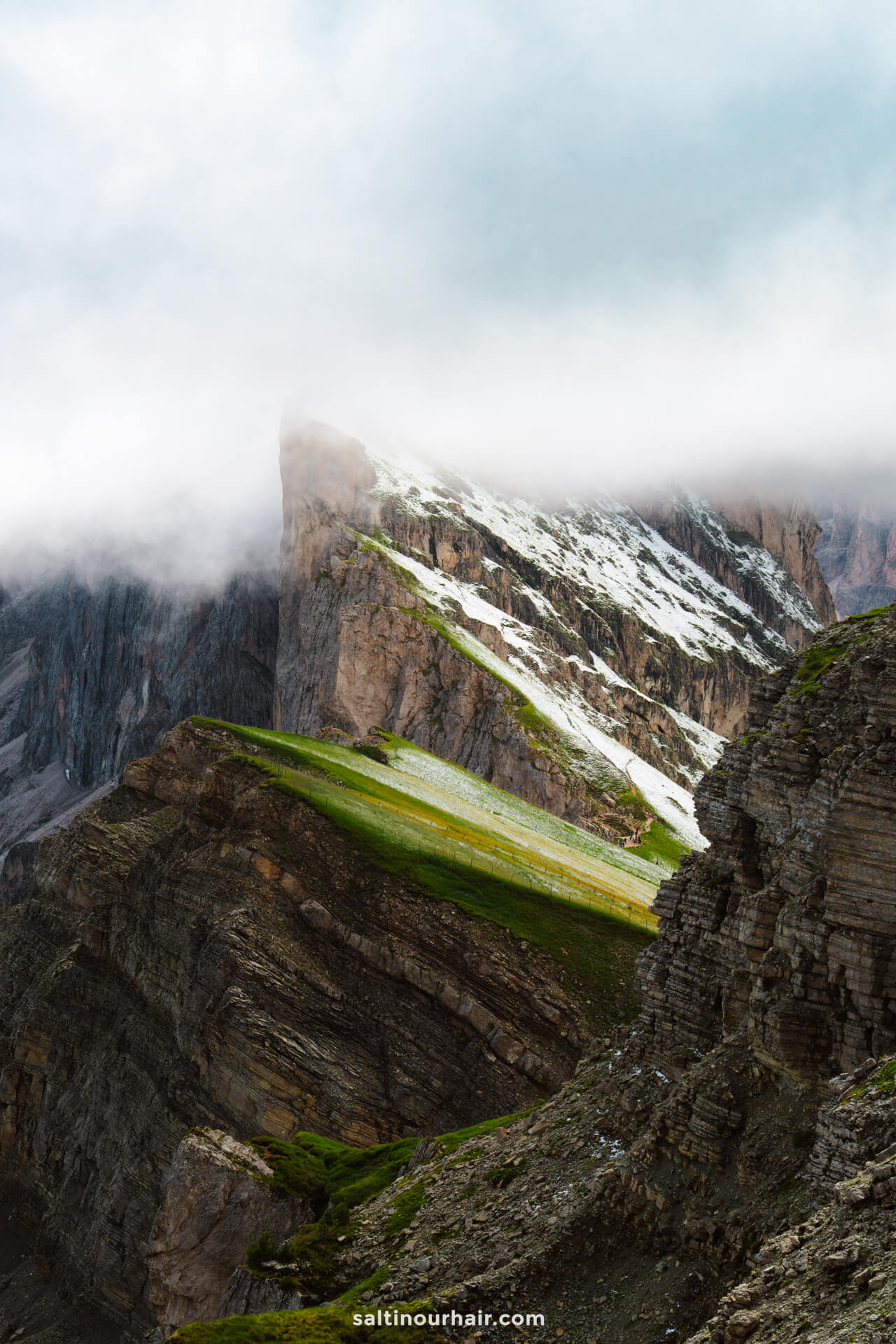 A rocky mountain landscape with patches of green grass and snow, partially covered by low-hanging clouds, frames the dramatic Seceda ridgeline in the dolomites of italy