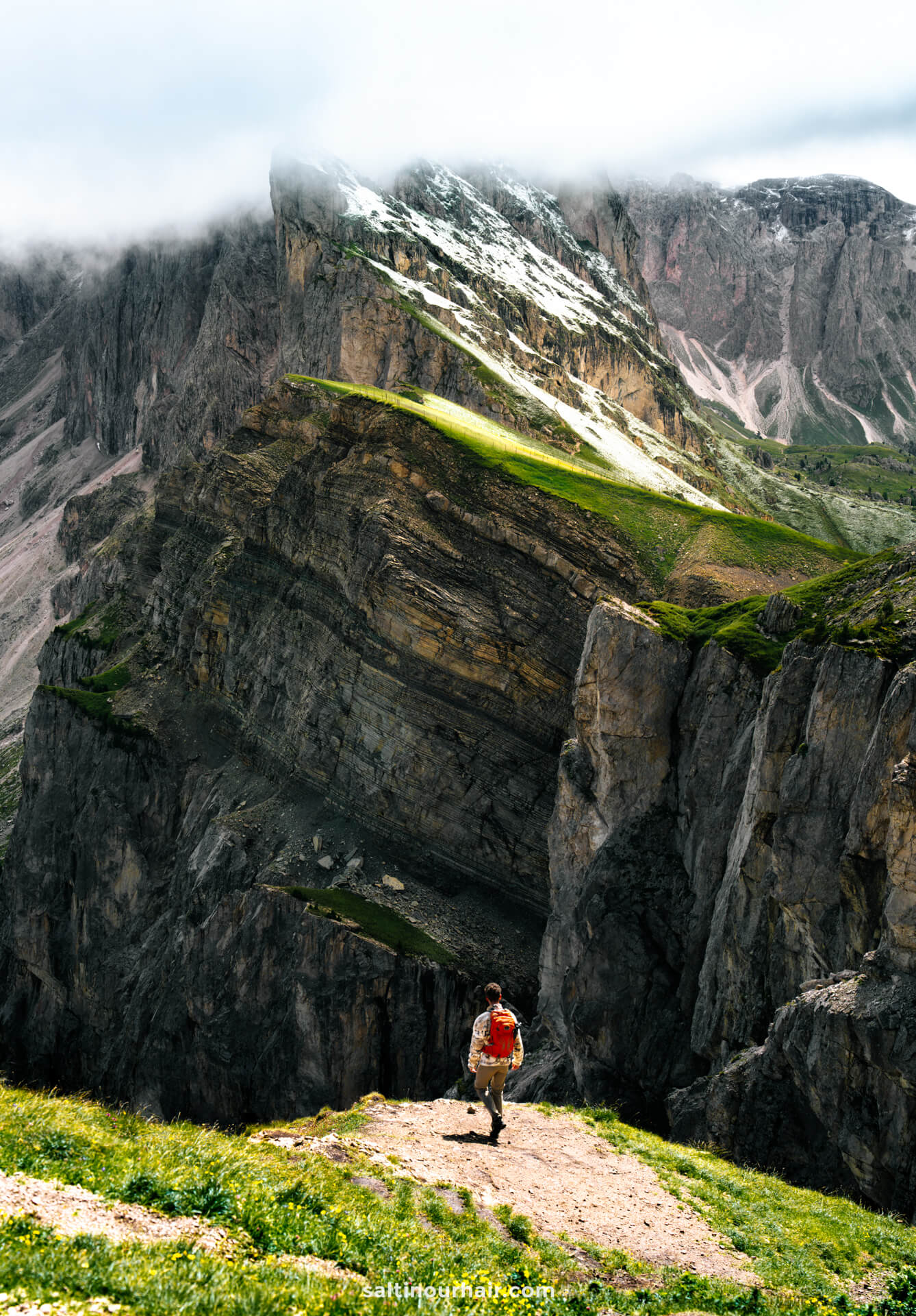 A man with a backpack stands on a grassy cliff edge along the Seceda ridgeline, facing steep rocky mountains with patches of snow and low clouds in the italian dolomites
