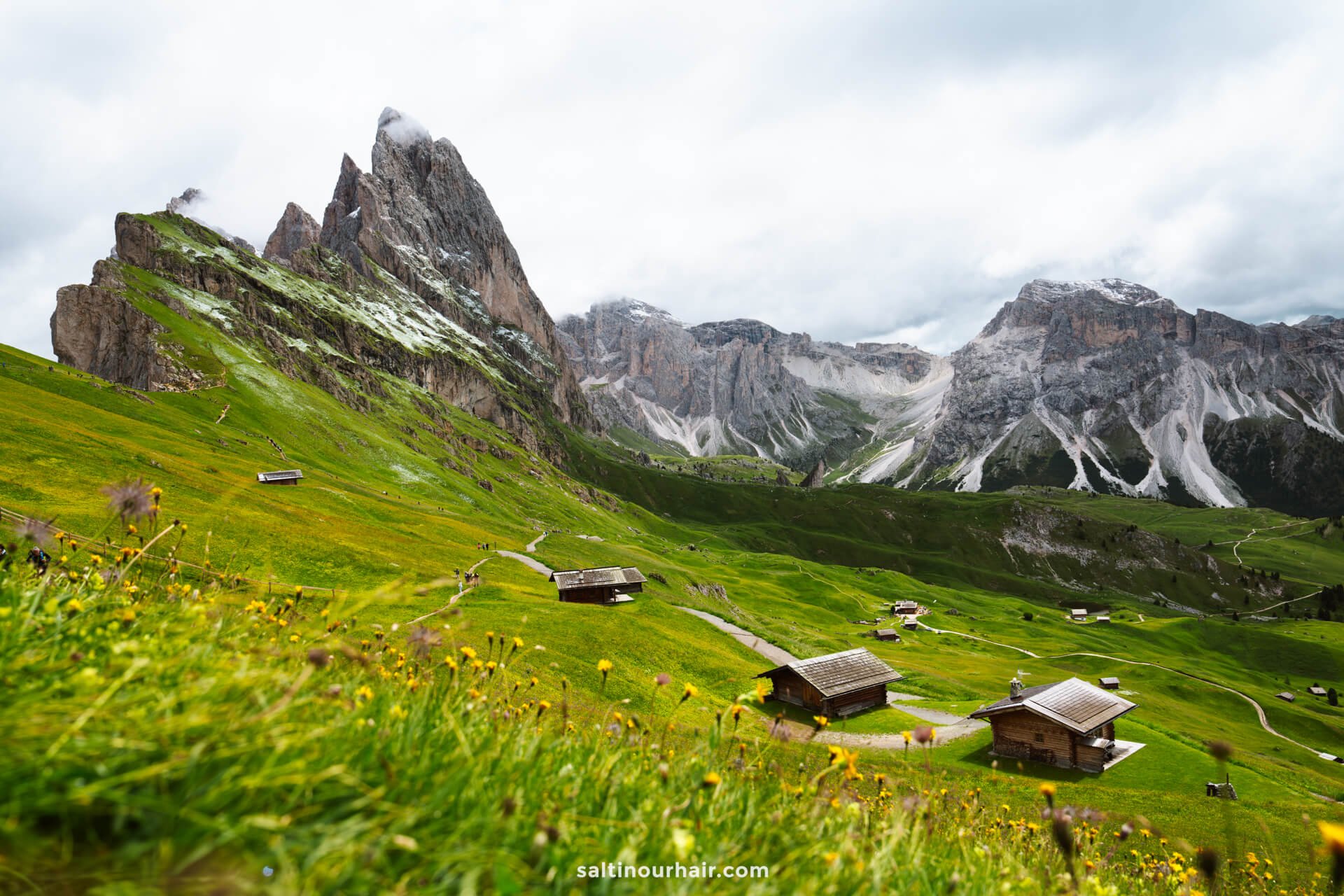 Wooden cabins are scattered on a grassy hillside with yellow flowers, set against the dramatic rocky peaks of the Seceda ridgeline and cloudy skies - a typical dolomites landscape