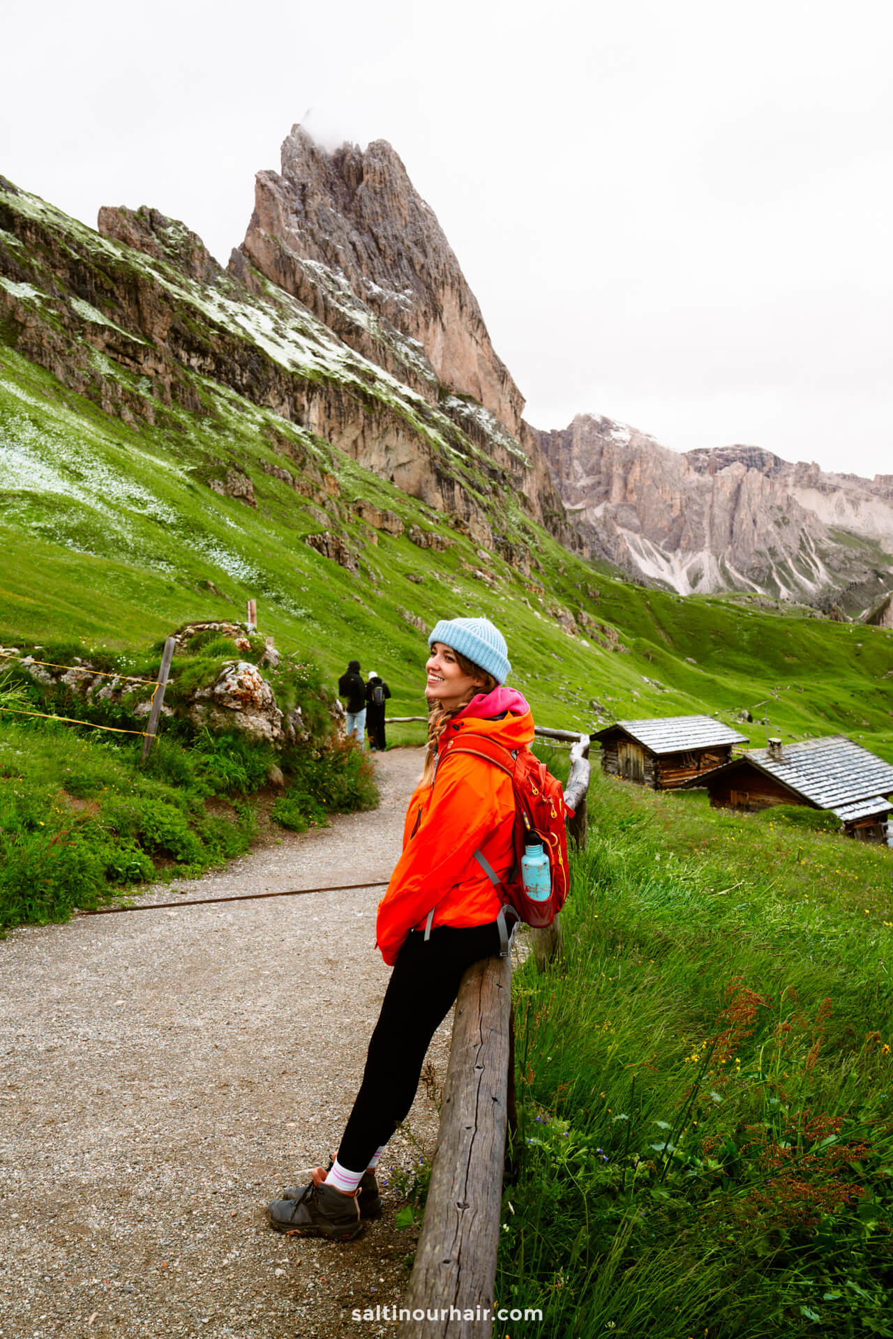 A woman in a red jacket and blue hat sits on a wooden fence along a mountain trail, with the stunning Seceda ridgeline and rocky peaks rising behind lush green hills of the dolomites italy
